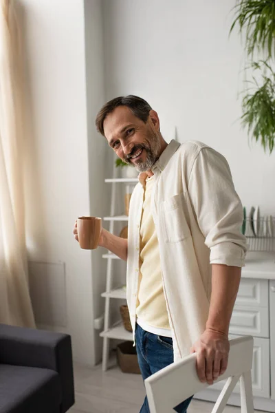 Joyeux homme barbu avec tasse de thé souriant à la caméra dans la cuisine — Photo de stock