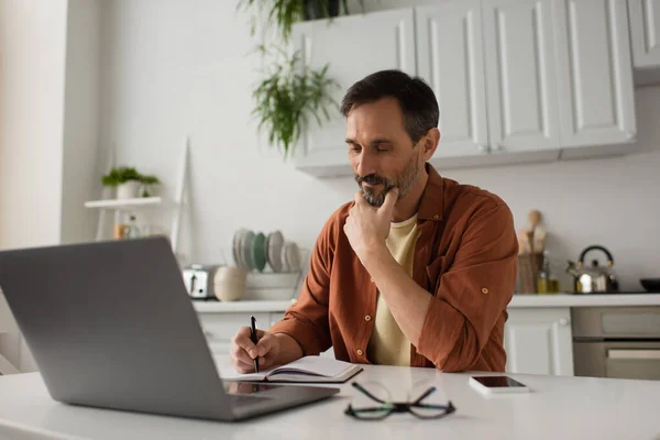 Uomo premuroso che tiene la mano vicino al viso e scrive nel notebook vicino al laptop in cucina — Foto stock