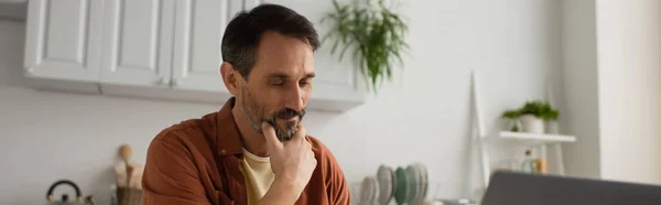 Hombre pensativo y sonriente tocando barba gris en cocina borrosa, pancarta - foto de stock