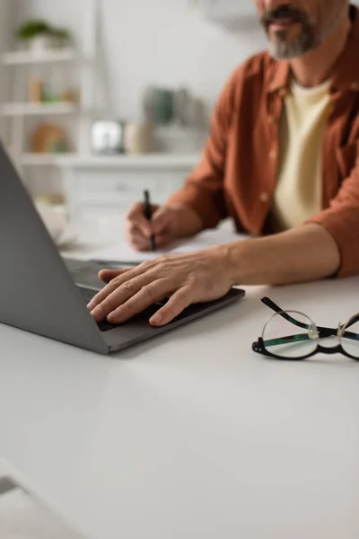 Partial view of blurred man working on laptop near eyeglasses on table — Stock Photo