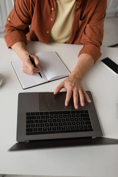 Вид сверху обрезанный человек с помощью ноутбука и записи в ноутбуке рядом с мобильным телефоном с пустым экраном — стоковое фото
