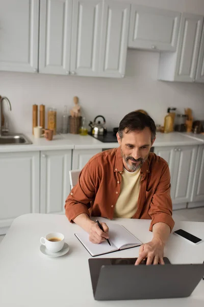 Homme souriant qui travaille avec un ordinateur portable et écrit dans un ordinateur portable près du café du matin et téléphone portable avec écran vierge — Photo de stock