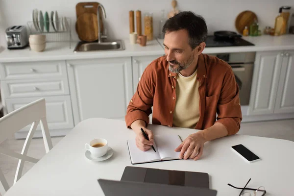 Бородатый человек пишет в блокноте возле ноутбука и кофейной чашки во время работы на кухне — стоковое фото