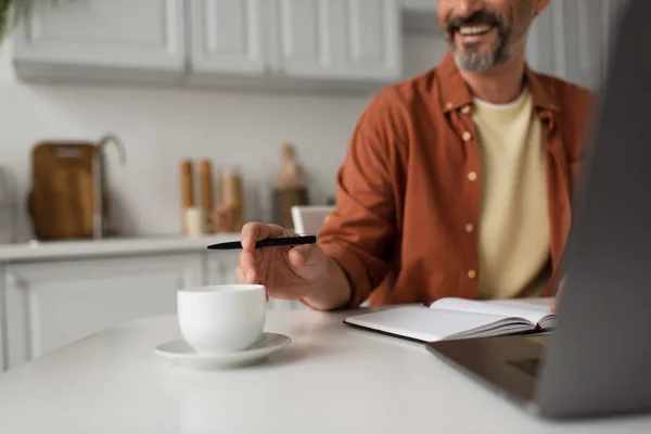 Vista recortada del hombre barbudo con pluma sonriendo cerca de la taza de café y el ordenador portátil borroso - foto de stock