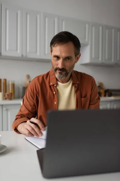 Homme réfléchi et positif regardant ordinateur portable flou tout en travaillant dans la cuisine — Photo de stock