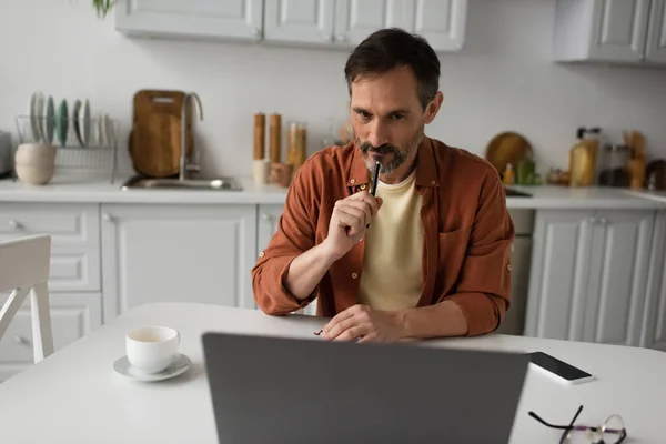 Cher homme avec stylo regardant ordinateur portable près tasse de café et smartphone avec écran blanc — Photo de stock