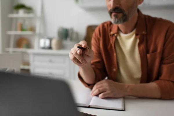 Ausgeschnittene Ansicht eines Mannes, der in der Küche arbeitet und mit Stift auf verschwommenen Laptop zeigt — Stockfoto