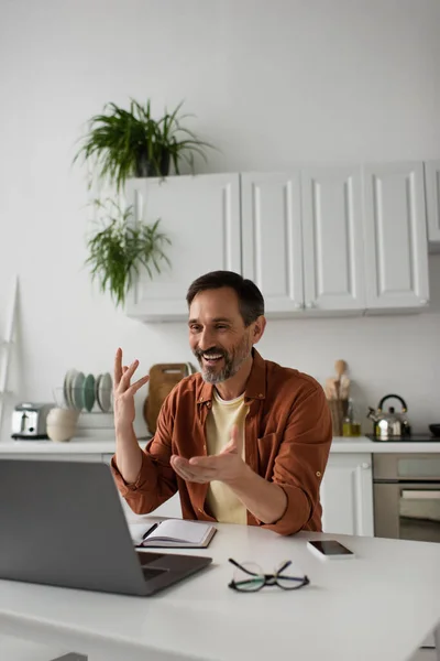 Cheerful man pointing at laptop during video call near smartphone and notebook in kitchen — Stock Photo