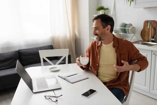 Felice uomo con tazza di caffè guardando computer portatile vicino notebook vuoto e smartphone con schermo bianco — Foto stock