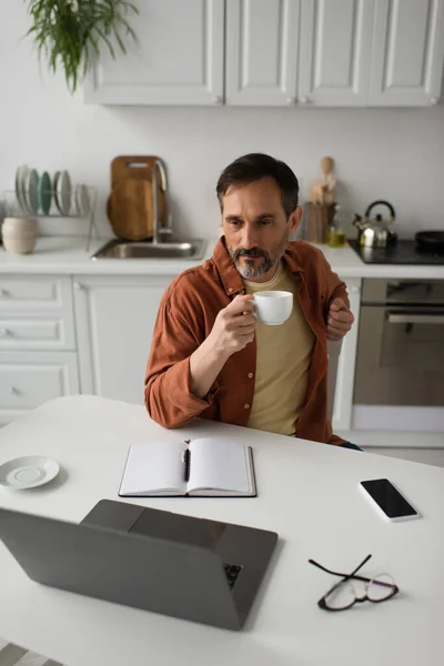 Hombre barbudo pensativo con taza de café mirando portátil cerca de portátil vacío y teléfono inteligente con pantalla en blanco - foto de stock