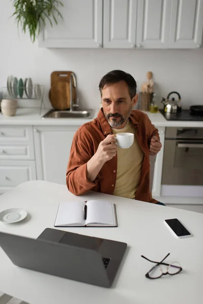 Pensive bearded man with coffee cup looking at laptop near empty notebook and smartphone with blank screen — Stock Photo