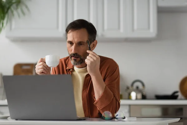 Barbudo hombre con taza de café tocando la mano con pluma mientras piensa cerca del ordenador portátil en la cocina - foto de stock
