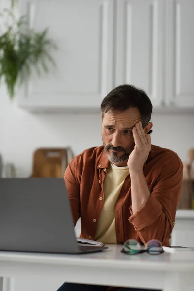 Homme fatigué regardant ordinateur portable flou dans la cuisine et souffrant de maux de tête — Photo de stock