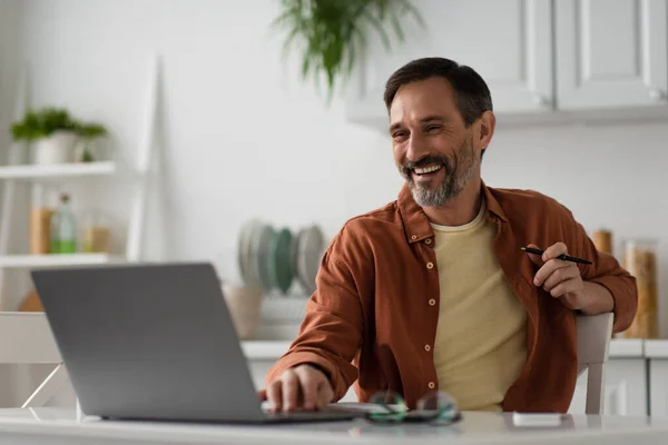 Щасливий чоловік працює на кухні і сміється, дивлячись на розмитий ноутбук — стокове фото