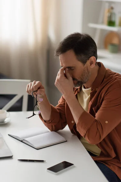 Uomo stanco che tiene gli occhiali e tocca gli occhi vicino al notebook vuoto e al cellulare con schermo vuoto — Foto stock