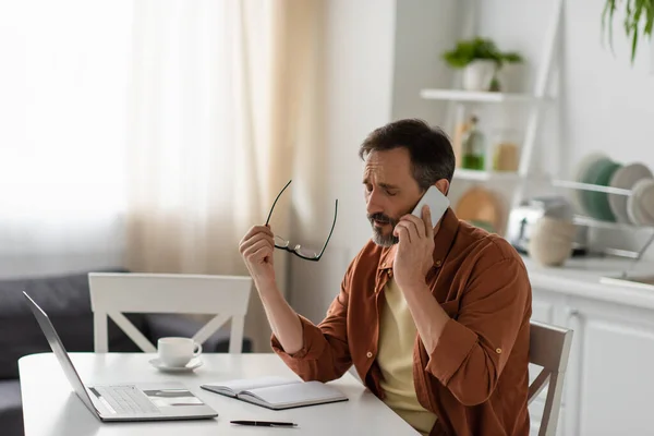 Frustrated man with closed eyes holding eyeglasses and talking on mobile phone near laptop — Stock Photo