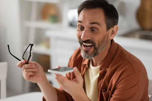 Astonished man holding eyeglasses while sending voice message on smartphone — Stock Photo