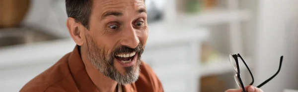 Homme barbu étonné tenant des lunettes à la maison sur fond flou, bannière — Photo de stock