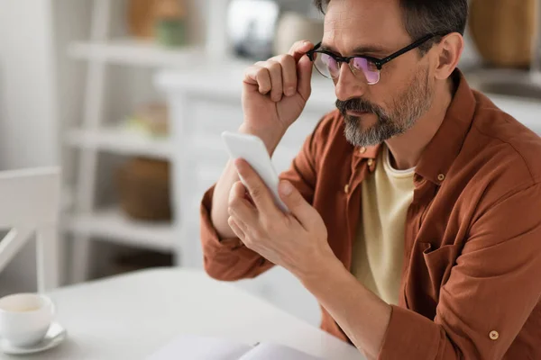 Bearded man adjusting eyeglasses and looking at mobile phone on blurred background — Stock Photo