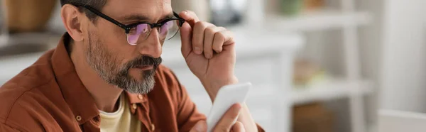 Hombre con barba gris mirando el teléfono inteligente mientras toca las gafas, pancarta - foto de stock