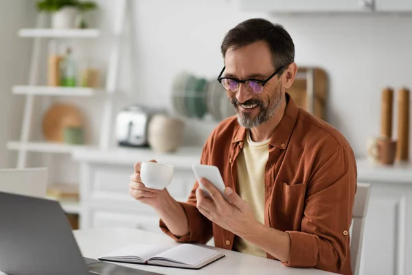 Uomo allegro con tazza di caffè guardando smartphone vicino notebook vuoto e laptop sfocato — Foto stock