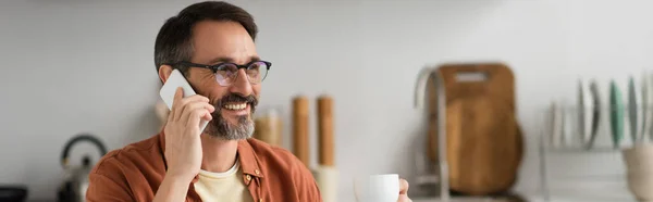 Hombre sonriente en anteojos sosteniendo taza de café y hablando por teléfono celular en la cocina, pancarta - foto de stock