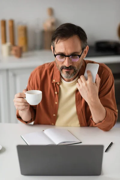 Mann mit Brille hält Smartphone und Kaffeetasse in der Hand und blickt auf verschwommenen Laptop neben leerem Notizbuch — Stockfoto