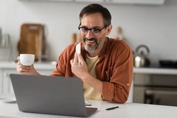 Улыбающийся мужчина в очках, держащий в руках мобильный телефон и кофейную чашку, глядя на размытый ноутбук — стоковое фото