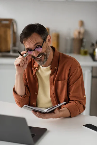 Веселий чоловік в окулярах тримає блокнот і ручку, посміхаючись на камеру біля розмитого ноутбука — стокове фото
