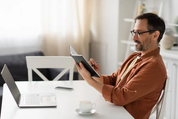 Seitenansicht eines lächelnden Mannes mit Brille, der in einem Notizbuch neben Laptop und Kaffeetasse schreibt — Stockfoto