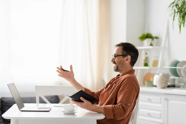 Side view of man in eyeglasses holding notebook and pointing with hand during video call on laptop — Stock Photo