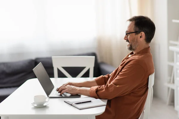 Side view of happy man in eyeglasses typing on laptop with blank screen near empty notebook and coffee cup — Stock Photo