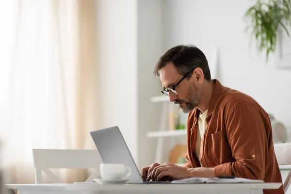 Focused man in eyeglasses typing on laptop near blurred coffee cup in kitchen — Stock Photo