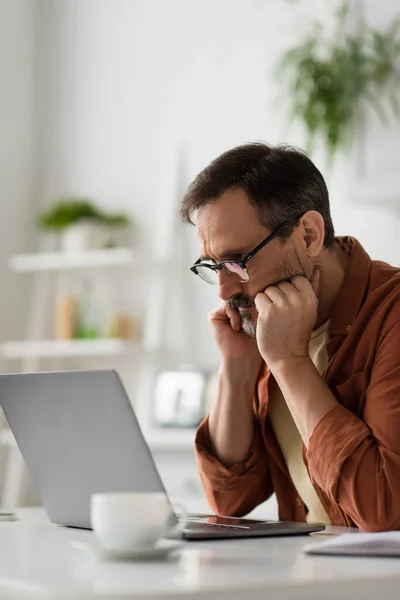 Cher homme dans les lunettes tenant la main près du visage tout en pensant à proximité d'un ordinateur portable dans la cuisine — Photo de stock