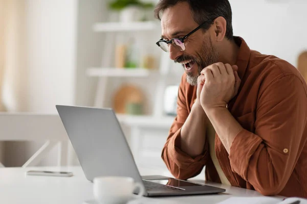 Amazed bearded man in eyeglasses looking at laptop while working in blurred kitchen — Stock Photo