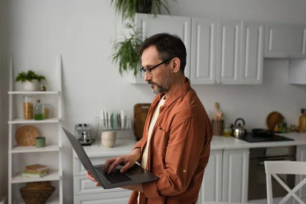 Vista lateral del hombre barbudo en gafas usando portátil mientras está de pie en la cocina - foto de stock