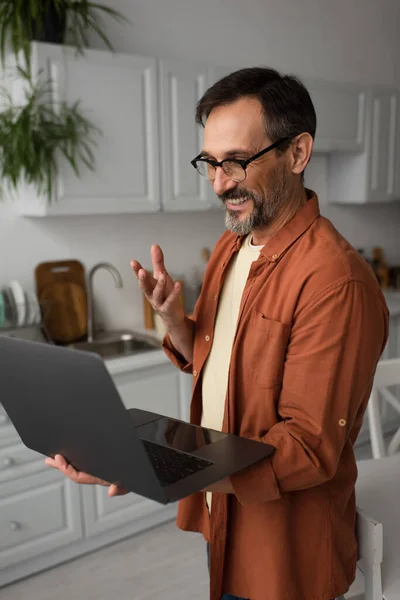 Щасливий бородатий чоловік в окулярах жестикулює під час відеодзвінка на ноутбук на кухні — стокове фото