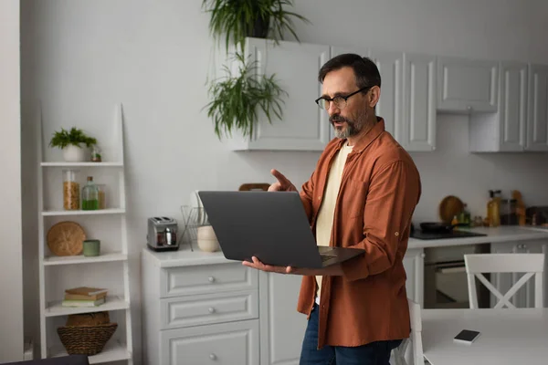 Homme en lunettes parler et pointant du doigt pendant le chat vidéo sur ordinateur portable dans la cuisine — Photo de stock