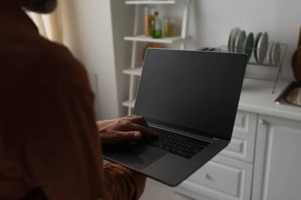 Vista parziale dell'uomo sfocato che tiene il computer portatile con schermo vuoto in cucina — Foto stock