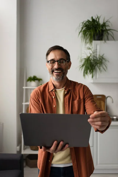 Excited man in eyeglasses holding laptop and laughing with closed eyes in kitchen — Stock Photo