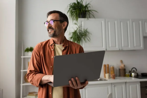Усміхнений чоловік в окулярах тримає ноутбук і дивиться на кухню — стокове фото