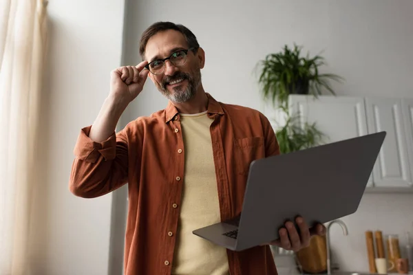 Bearded man adjusting eyeglasses and holding laptop while smiling at camera — Stock Photo