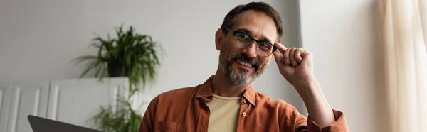 Cheerful bearded man adjusting eyeglasses while smiling at camera in kitchen, banner — Stock Photo