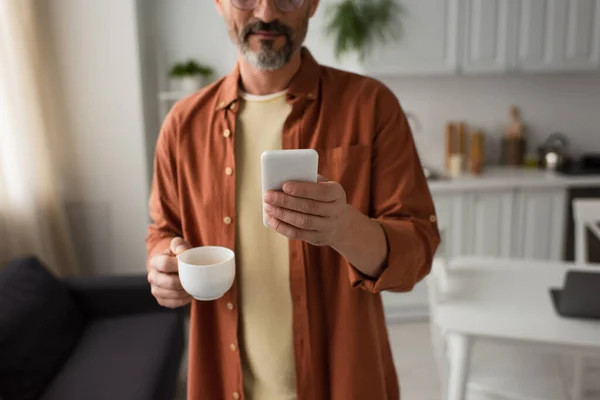 Vista cortada do homem barbudo na camisa marrom segurando smartphone e xícara de café na cozinha — Fotografia de Stock