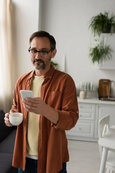 Uomo in occhiali e camicia marrone con tazza di caffè e messaggistica sul cellulare in cucina — Foto stock