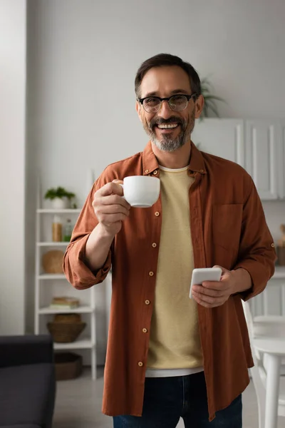 Felice uomo in occhiali in possesso di tazza di caffè e telefono cellulare mentre guardando la fotocamera in cucina — Foto stock