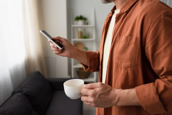 Vue partielle de l'homme en chemise brune tenant une tasse de café blanche et utilisant un téléphone mobile à la maison — Photo de stock