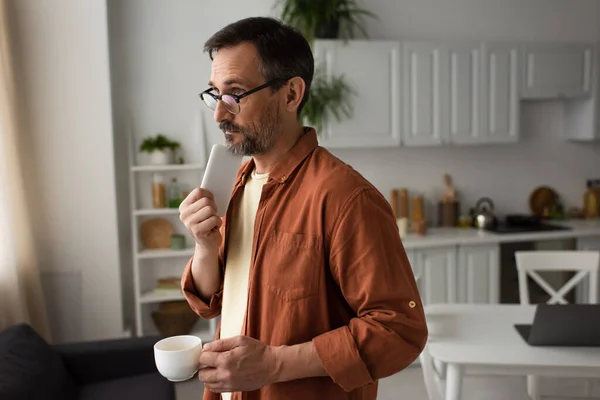 Hombre reflexivo en gafas de pie con teléfono inteligente y taza de café cerca del ordenador portátil sobre fondo borroso - foto de stock