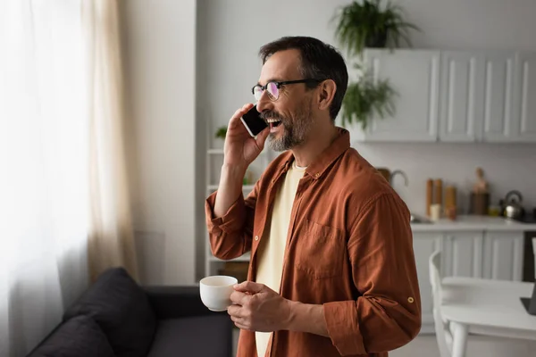 Man in brown shirt and eyeglasses holding coffee cup and talking on smartphone in kitchen — Stock Photo