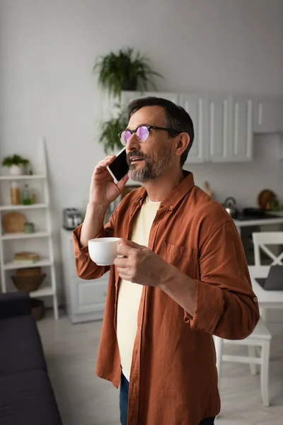 Бородатий чоловік в окулярах говорить на смартфоні і тримає чашку кави на кухні — стокове фото
